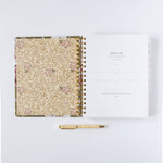 2026 Meadow weekly planner with floral cover, open to the first page, and a gold pen beside it on a white background.