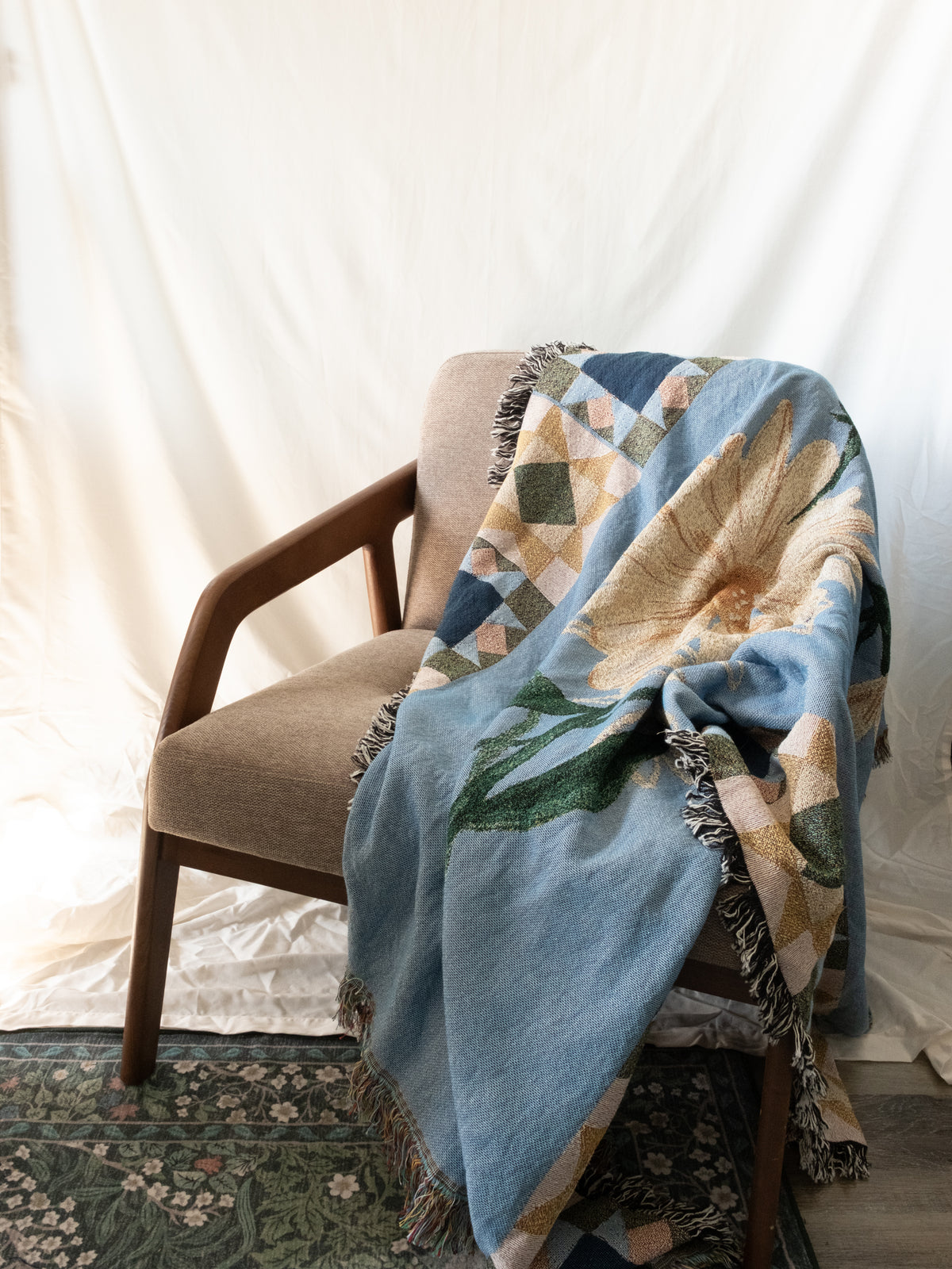 Blue woven fabric blanket with a yellow flower and green leaves draped over a wooden chair with a tan cushion.
