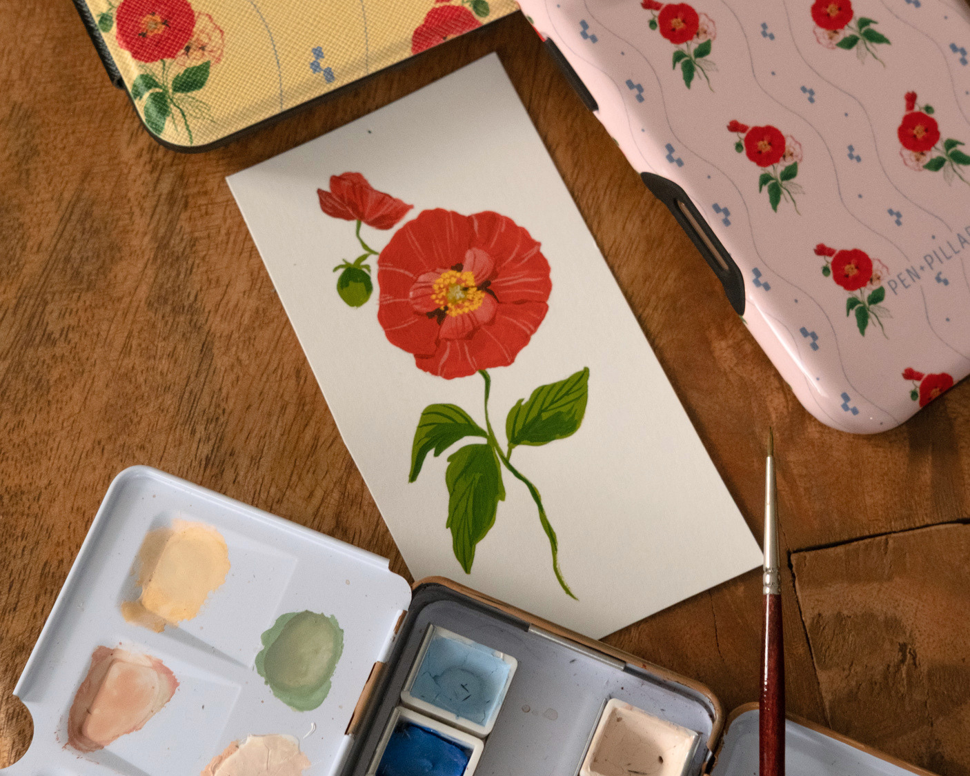 Yellow floral Kindle case and pink phone case by PEN+PILLAR next to a watercolor painting and paint set on a wood table.