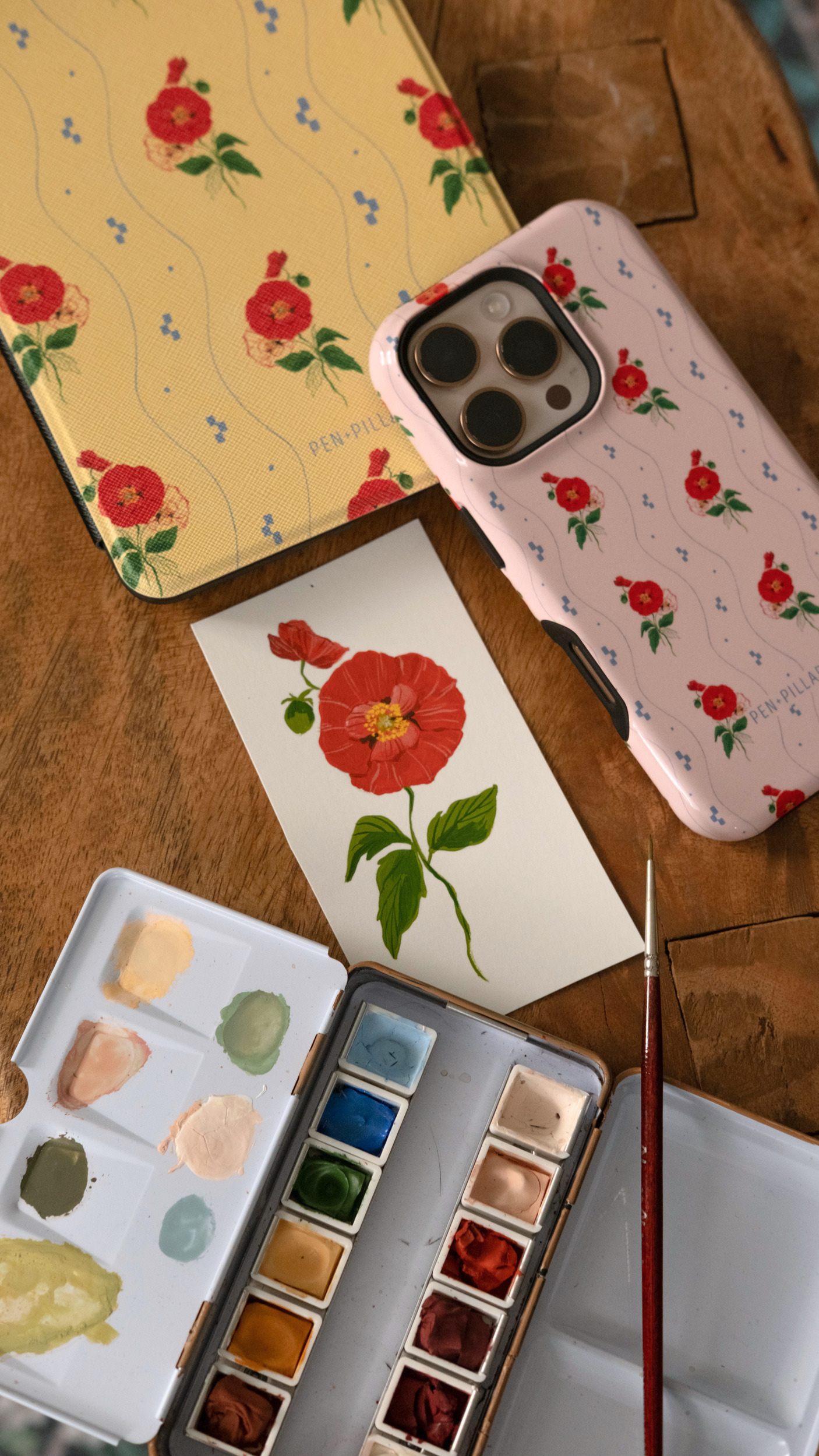 Pink plastic iPhone case with red poppy pattern next to a watercolor painting, palette, and brush on a wooden surface.