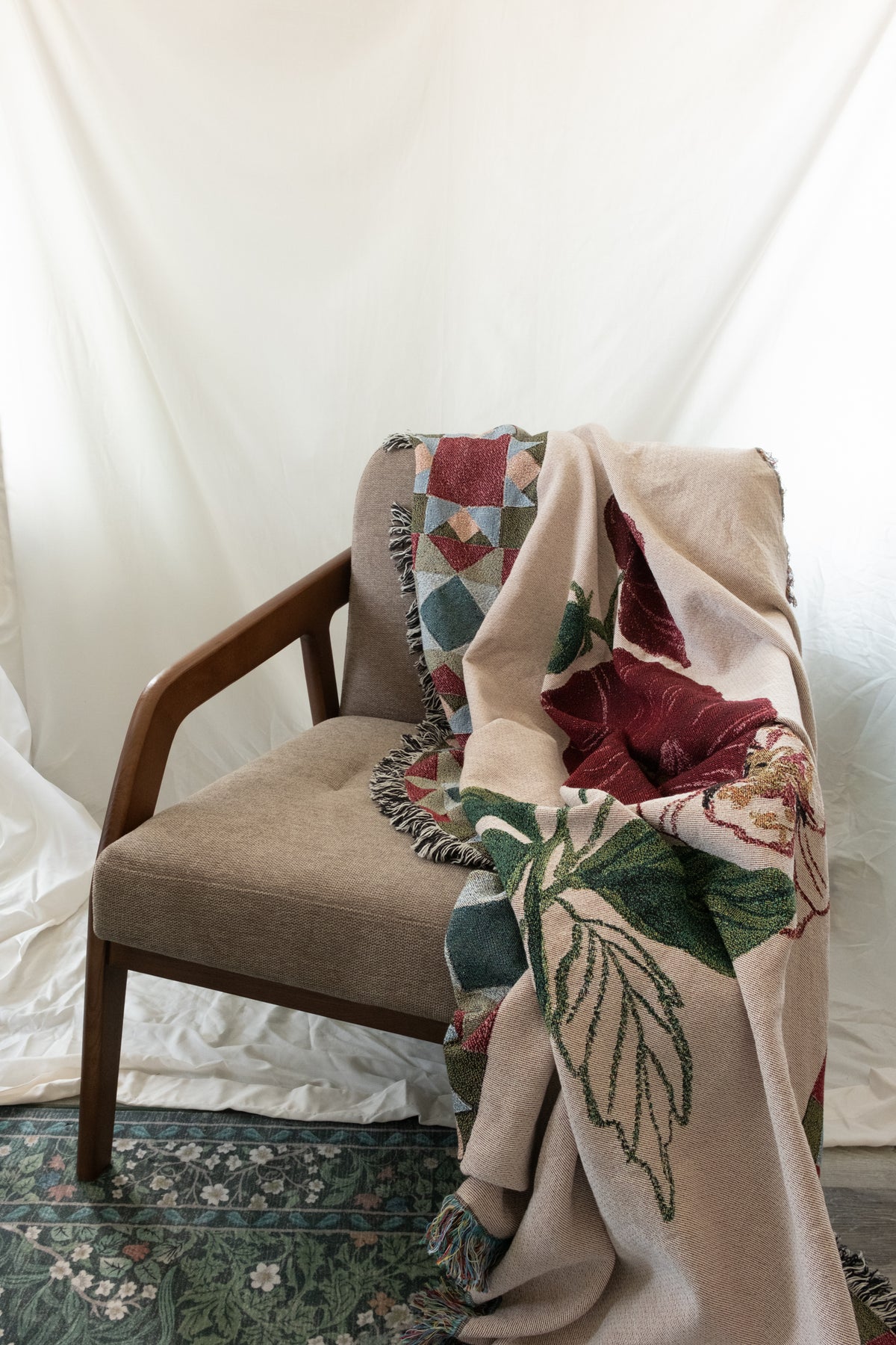 Woven fabric blanket with red floral and green leaf patterns draped over a brown fabric and wood chair.