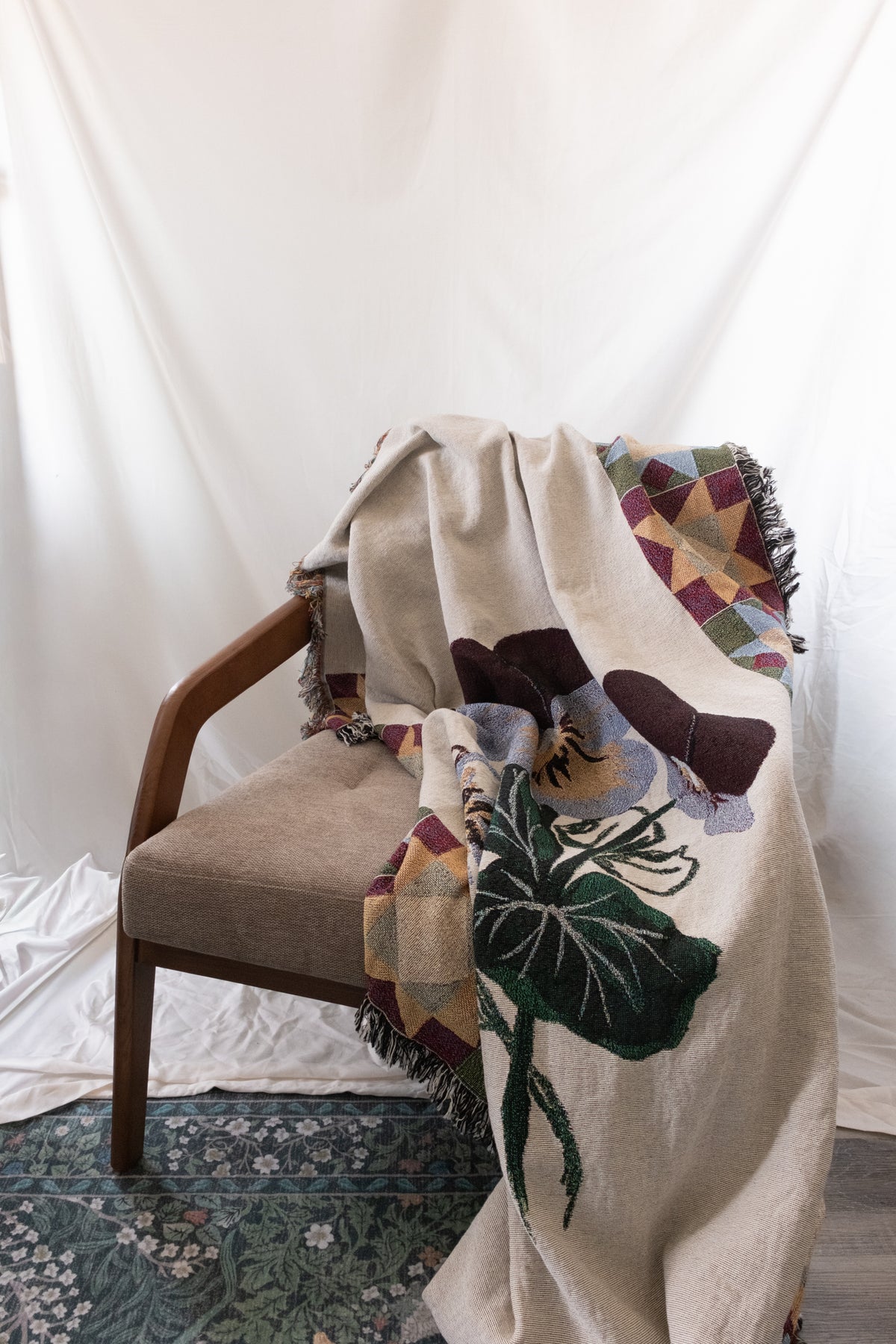 Cream woven fabric blanket with purple flowers and green leaves draped over a wooden chair with a fabric cushion.
