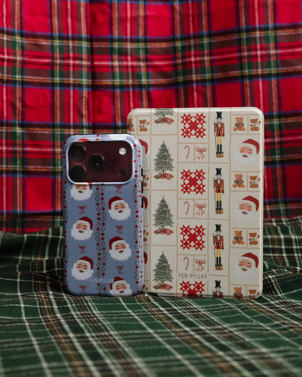 Holiday Sampler Kindle case standing beside a Jolly phone case on red plaid