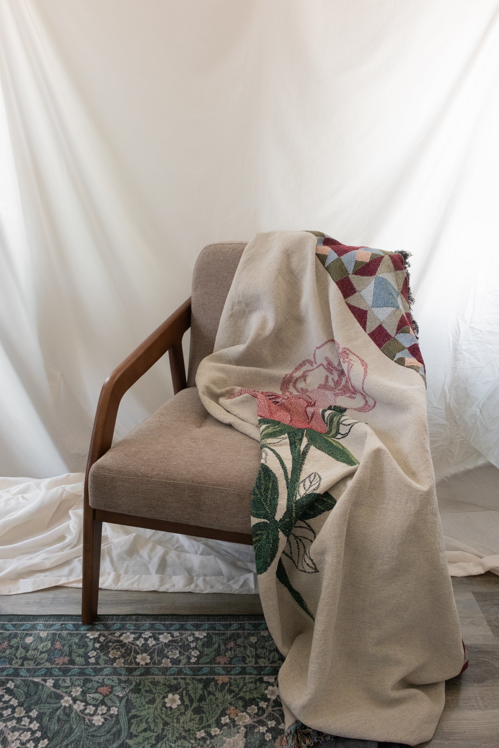 Cream woven fabric blanket with a pink rose and green leaves draped over a brown fabric and wood chair.