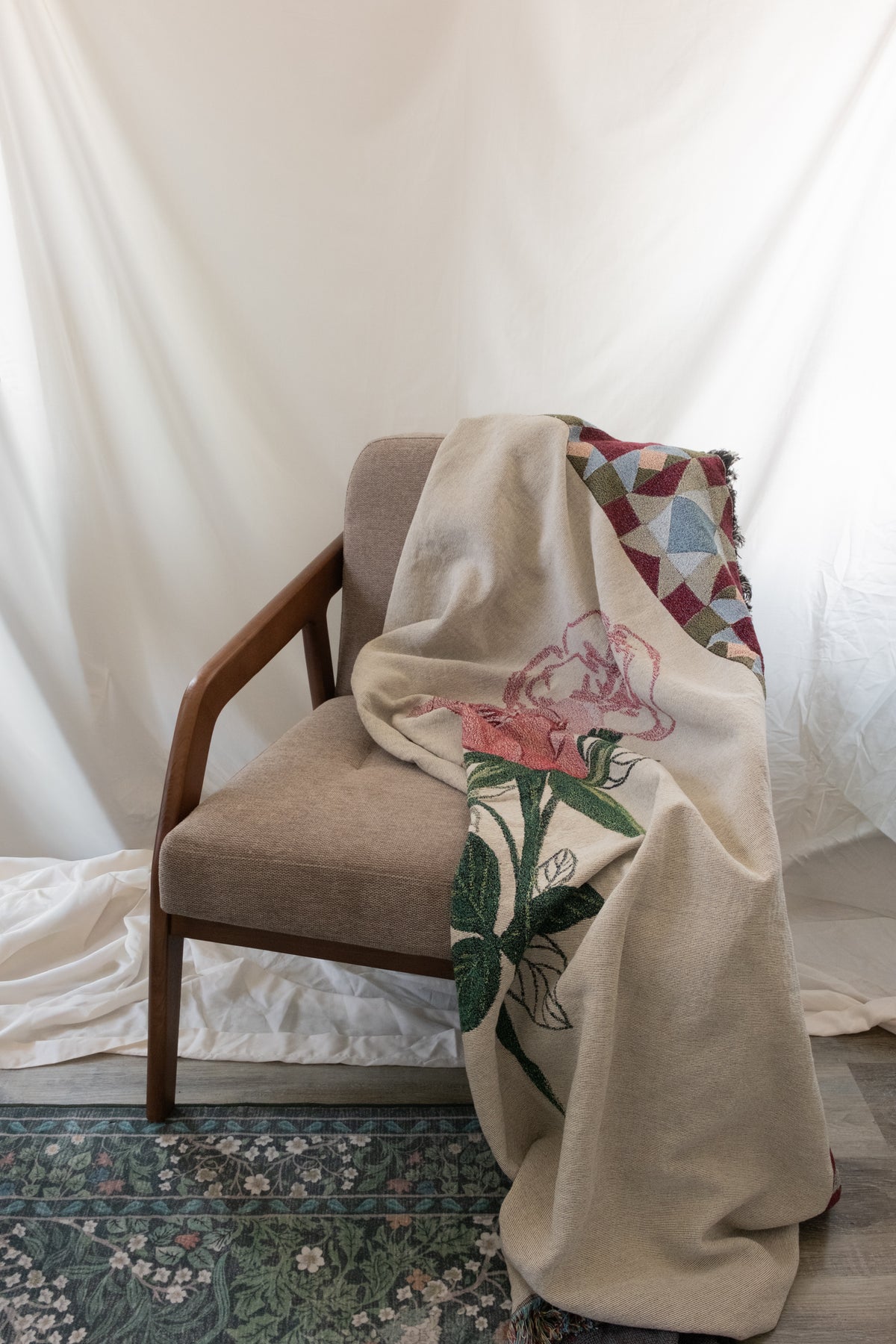 Cream woven fabric blanket with a pink rose and green leaves draped over a brown fabric and wood chair.