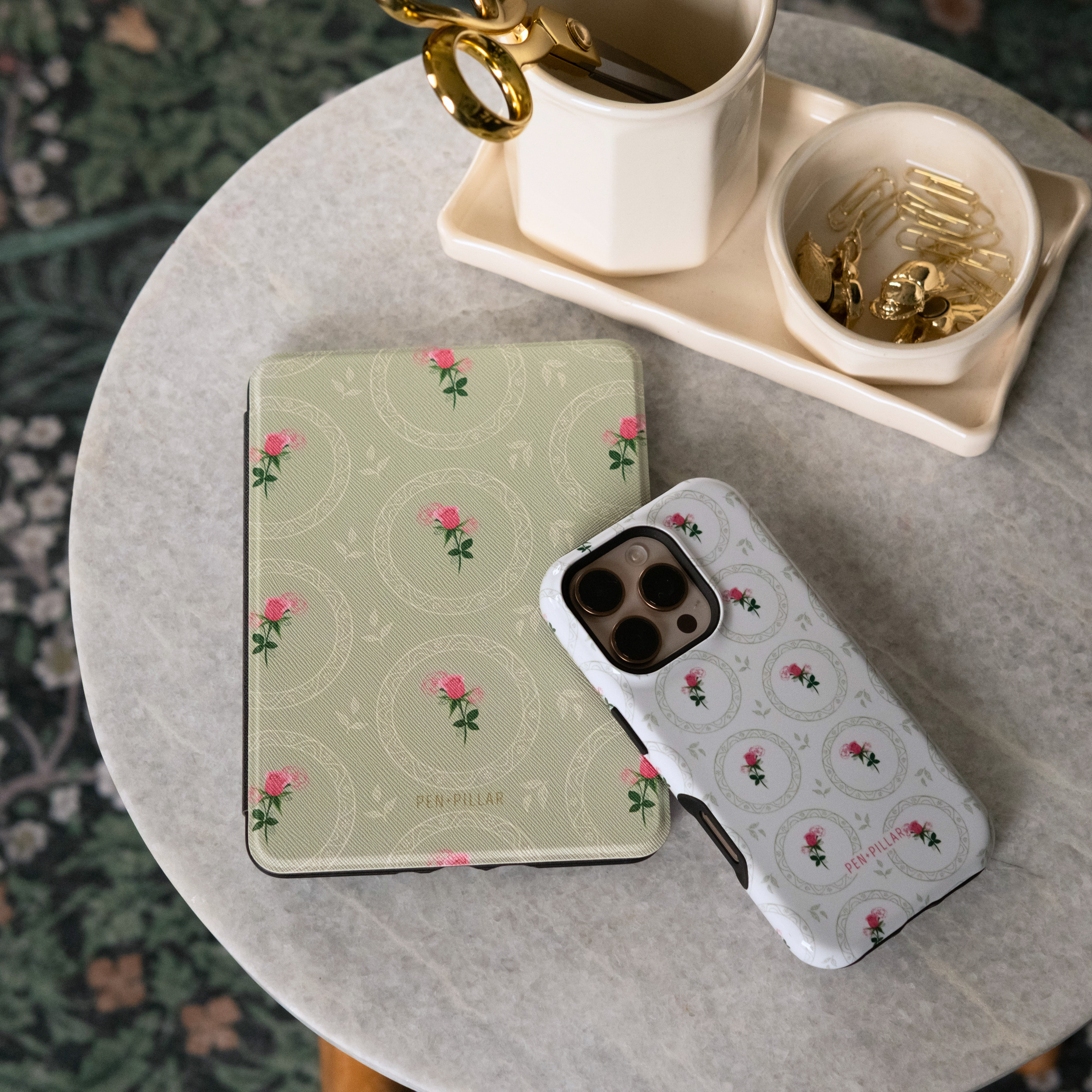 Sage green Kindle case and white phone case with pink rose patterns by PEN+PILLAR on a stone table.