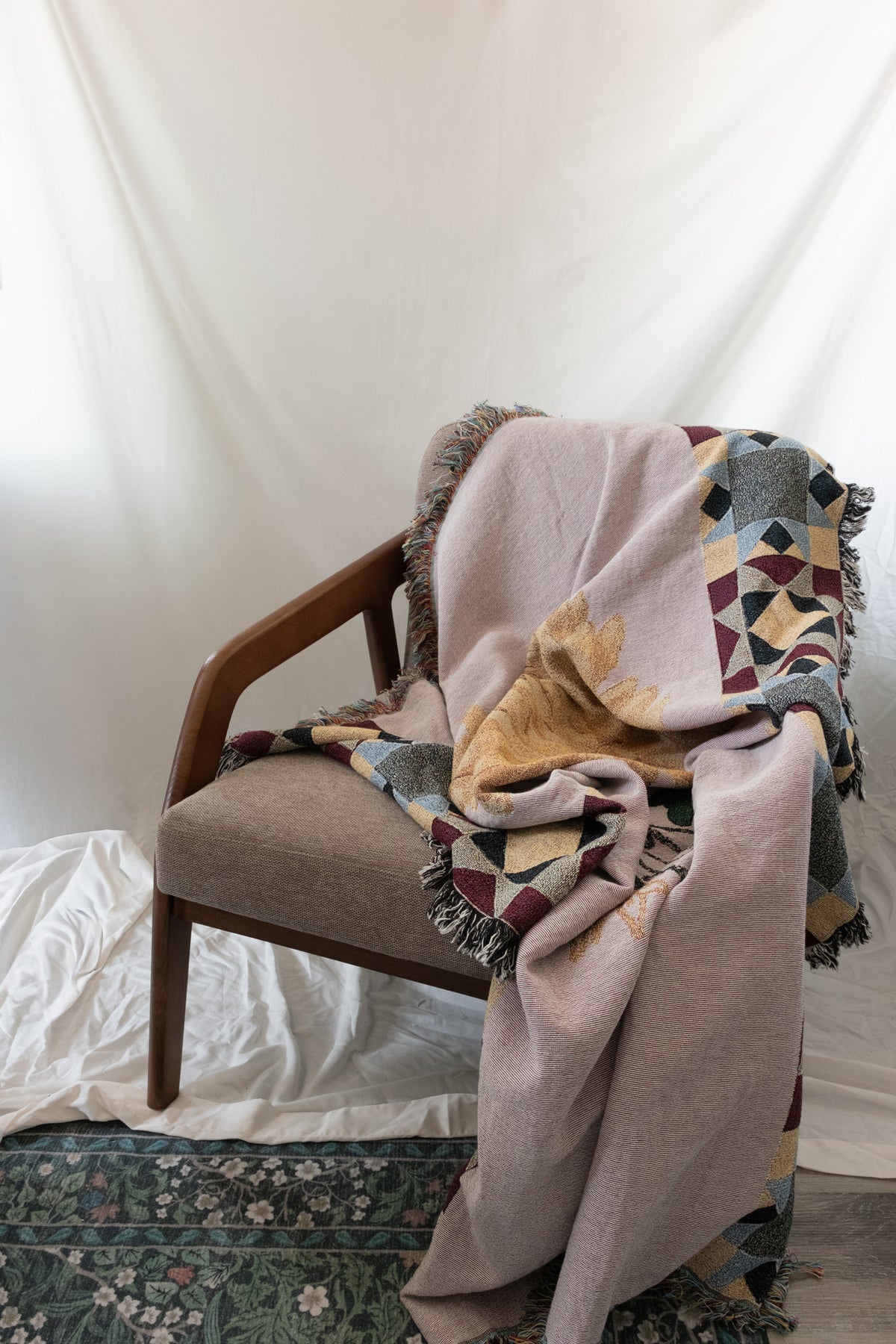 Woven pink blanket with geometric borders and fringe draped over a wooden armchair on a white fabric backdrop.