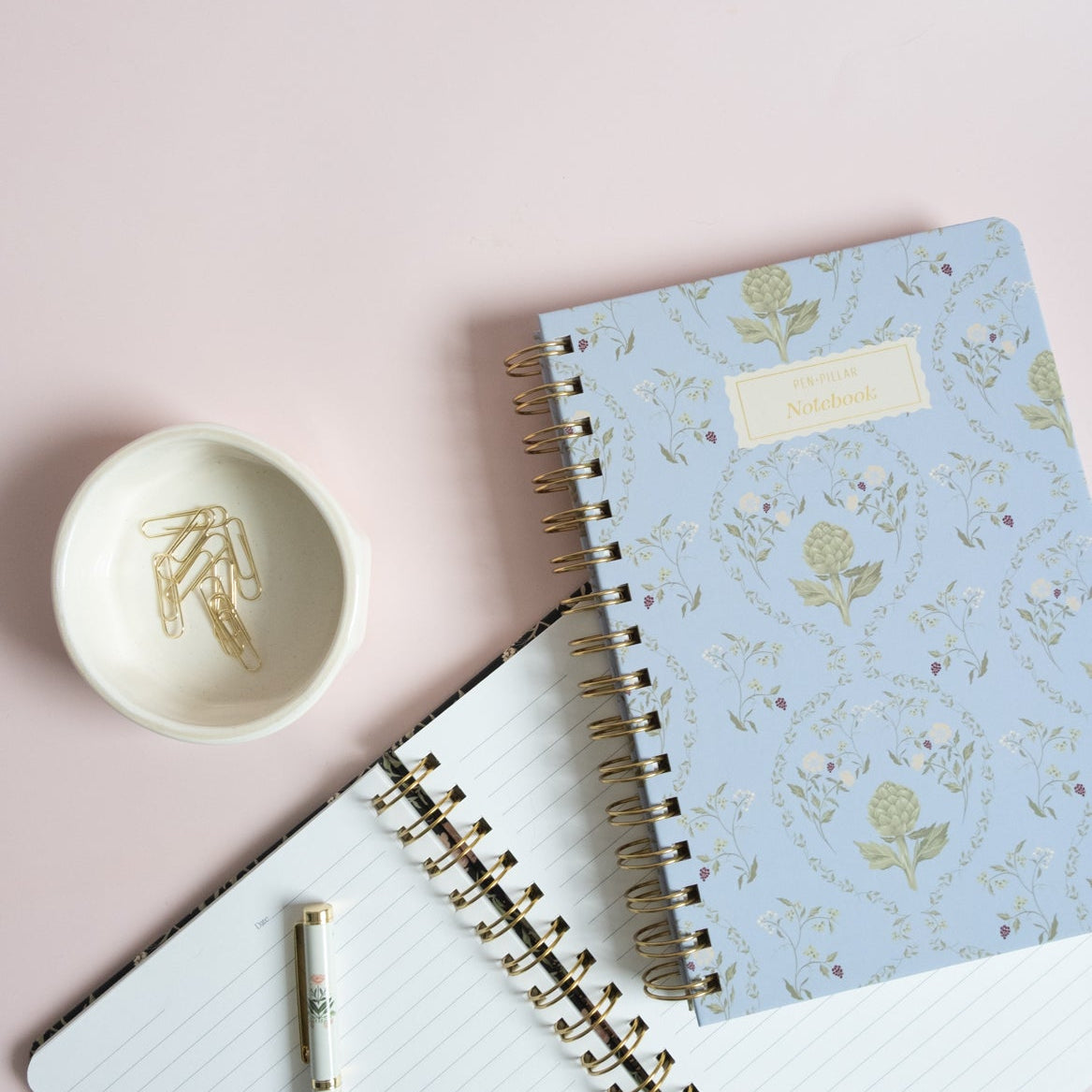 Artichoke notebook with a light blue floral cover, placed on a pink background with a white spiral notebook and a small bo...