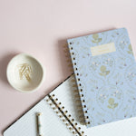 Artichoke notebook with a light blue floral cover, placed on a pink background with a white spiral notebook and a small bo...