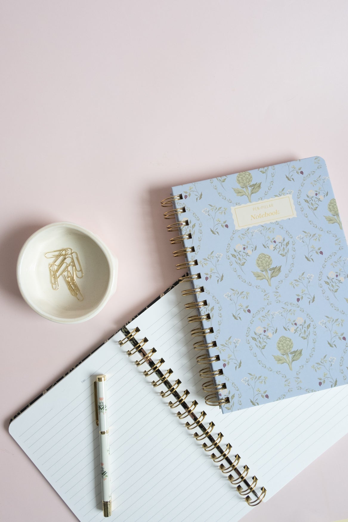 Artichoke notebook with a light blue floral cover, placed on a pink background with a white spiral notebook and a small bo...