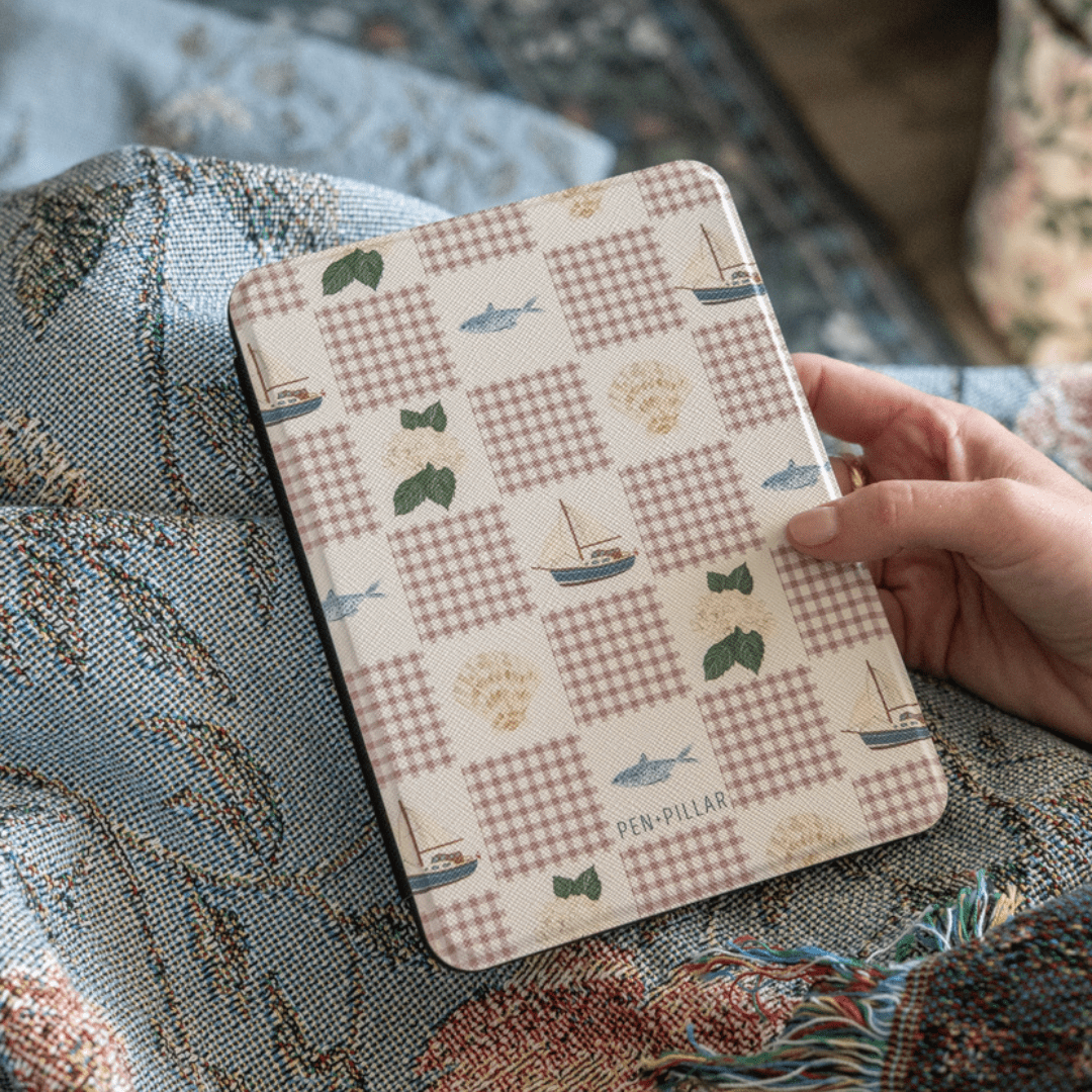 Rectangular PEN PILLAR Kindle case with a pink checkered pattern, sailboats, and fish held against a blue woven blanket.