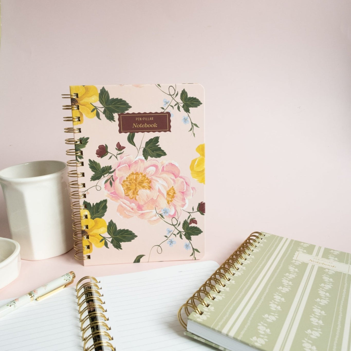 Garden rose notebook with floral design, surrounded by a white cup, a pen, and two other notebooks on a pink background.