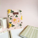 Garden rose notebook with floral design, surrounded by a white cup, a pen, and two other notebooks on a pink background.