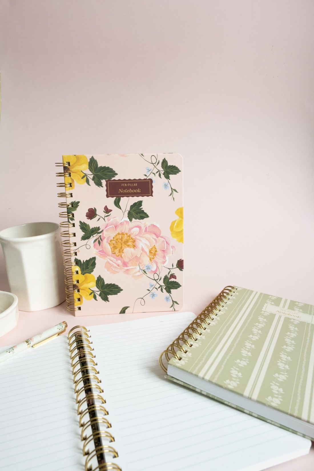 Garden rose notebook with floral design, surrounded by a white cup, a pen, and two other notebooks on a pink background.