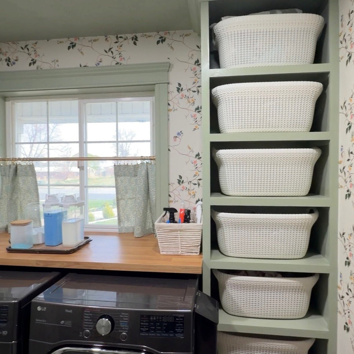Grove floral wallpaper with colorful birds and branches behind green shelving and white woven baskets in a laundry room.