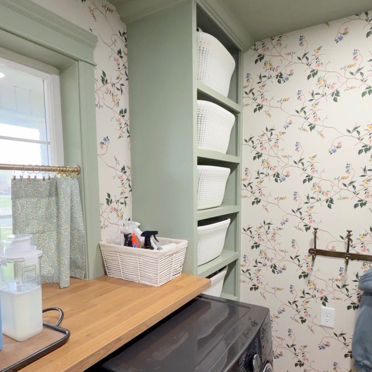 Grove floral wallpaper with pink and blue flowers on a white background in a laundry room with sage green shelving.