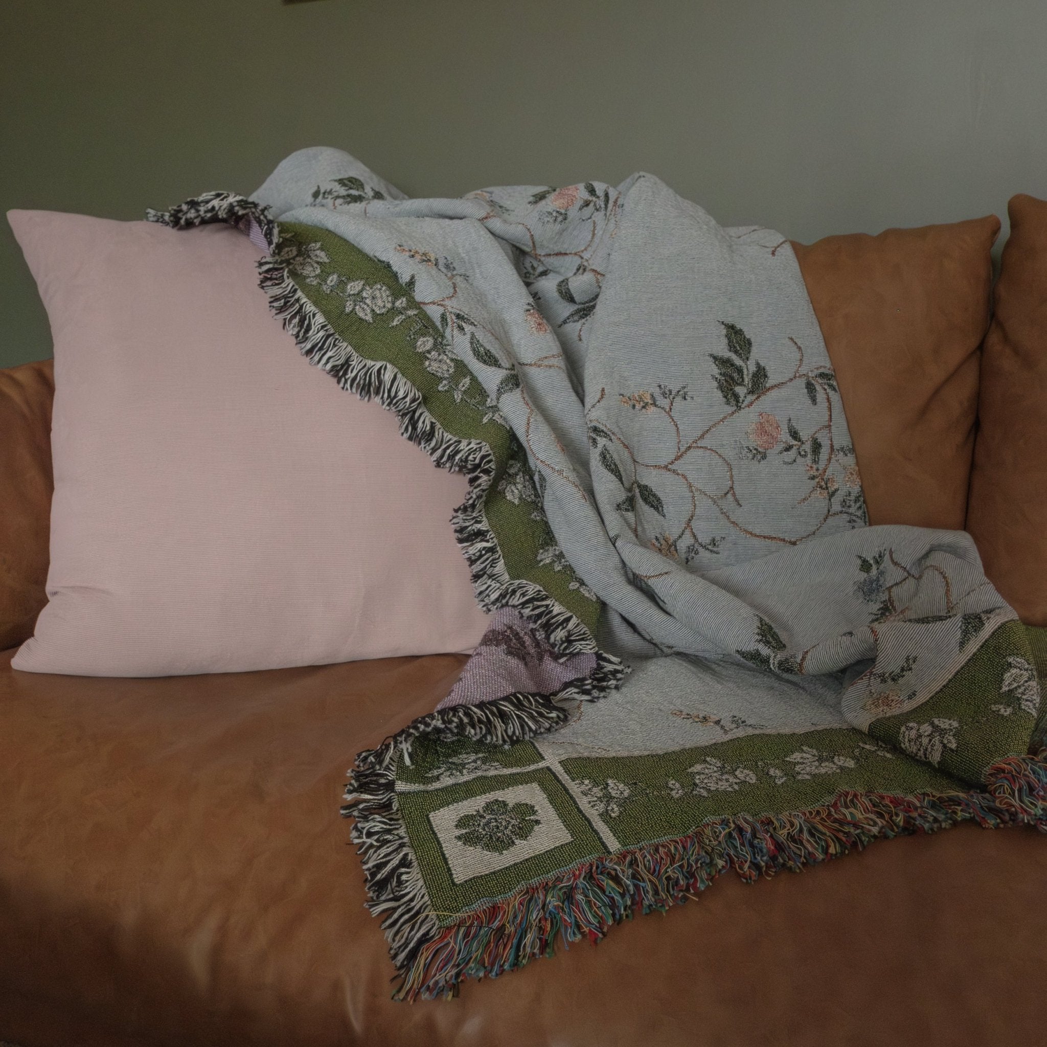 Grove woven blanket in light blue with floral patterns, draped over a brown sofa beside a pink pillow.