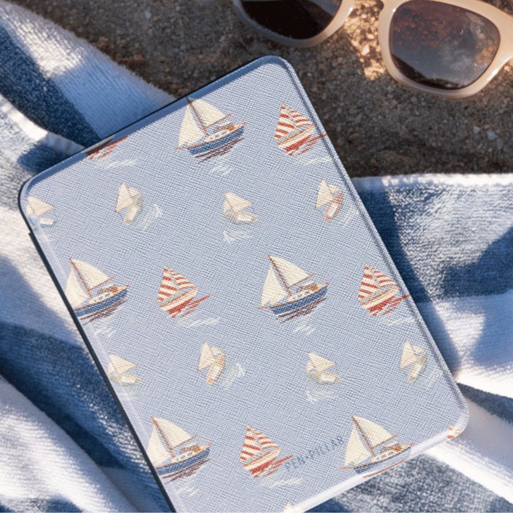 Light blue Harbor Kindle Case with a sailboat pattern on a beach towel next to sunglasses. The label reads PEN PILLAR.