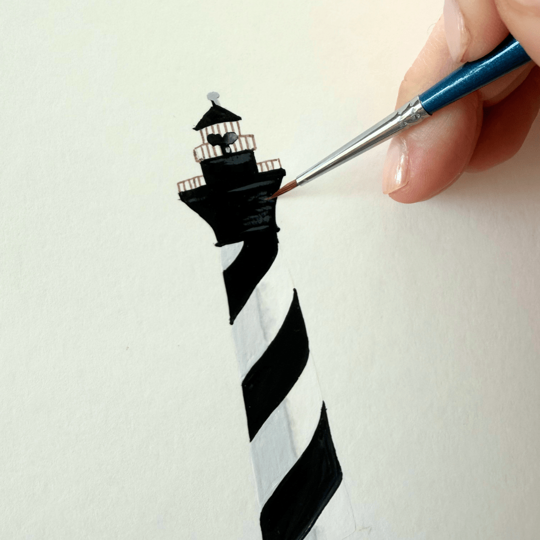 A hand uses a thin blue brush to paint a black and white spiraled lighthouse on a white surface.