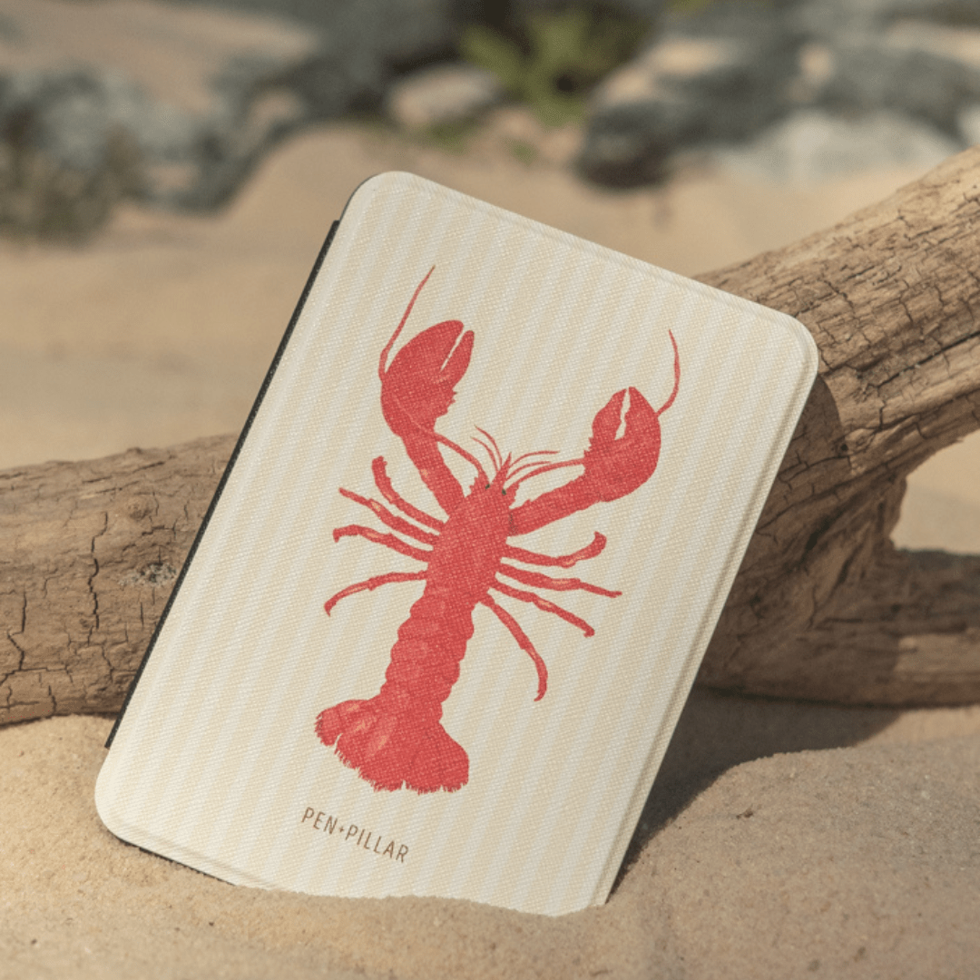 Cream Kindle case with a red lobster illustration and PEN+PILLAR text, resting on sand against a piece of wood.