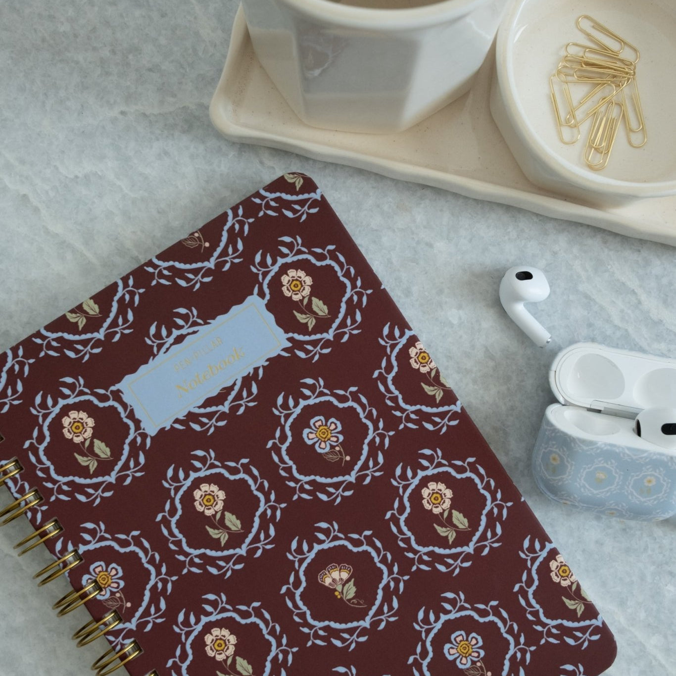 Merrigold notebook with floral design, gold spiral binding, placed on a marble surface with a cup and stationery.