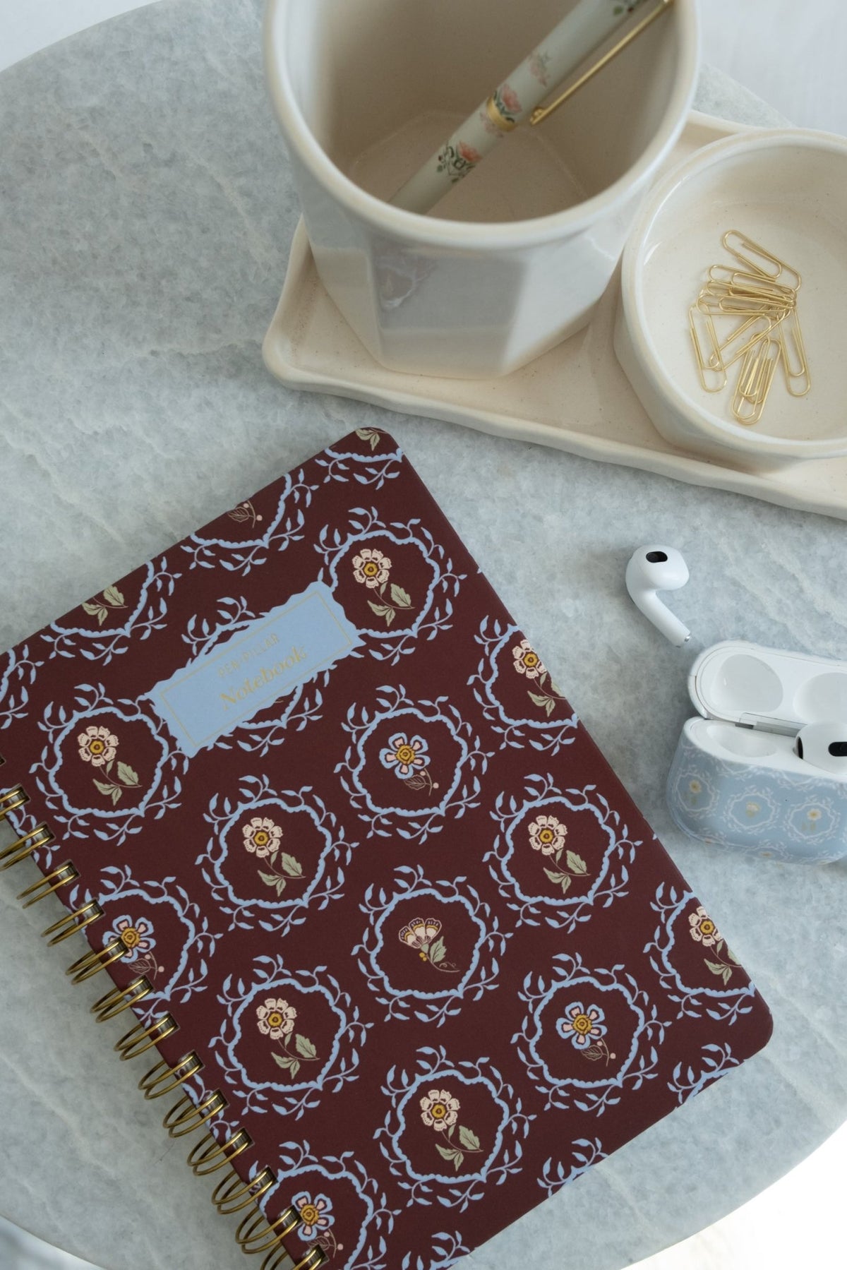 Merrigold notebook with floral design, gold spiral binding, placed on a marble surface with a cup and stationery.