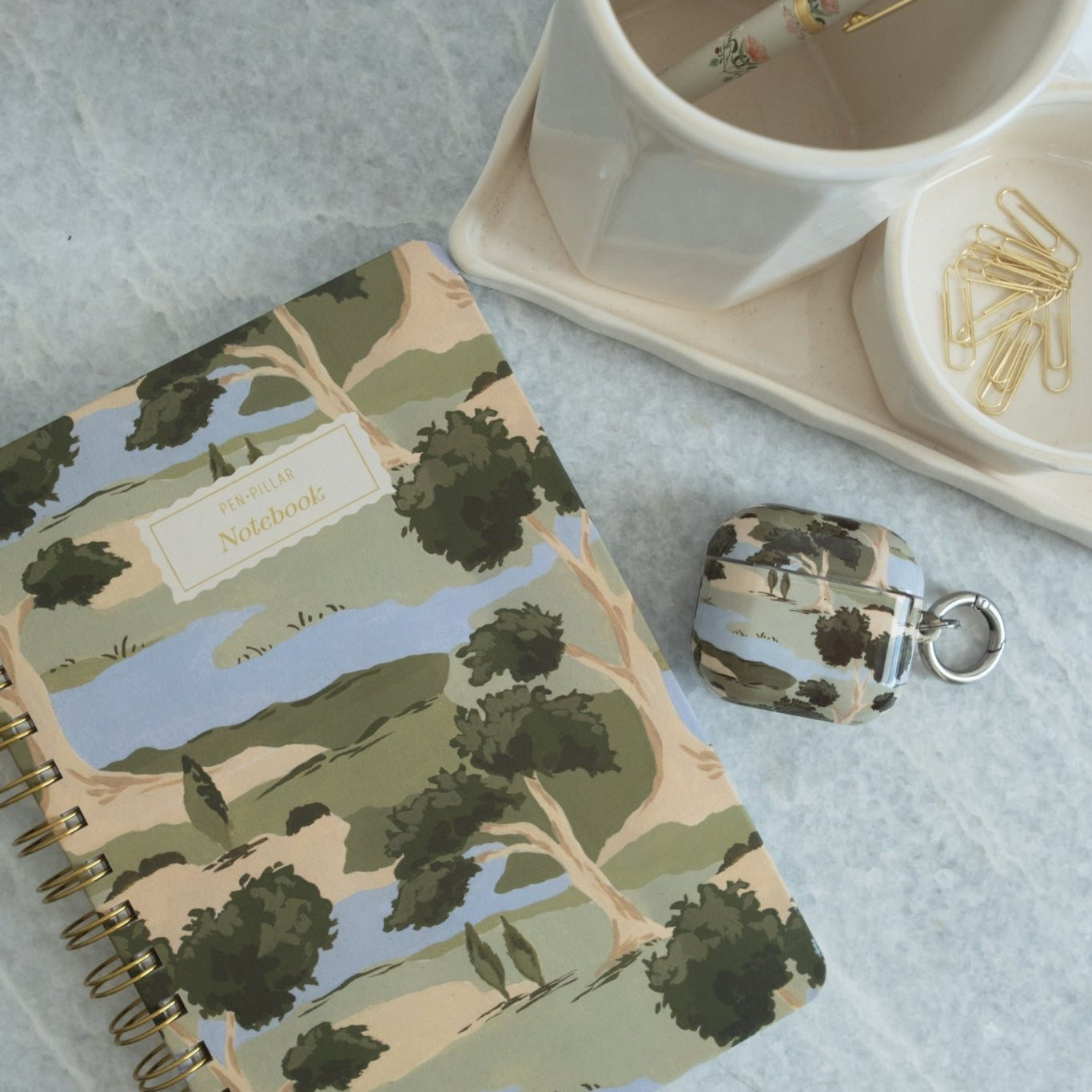Orchard notebook with a colorful landscape design, placed on a marble table beside a cup and paper clips.