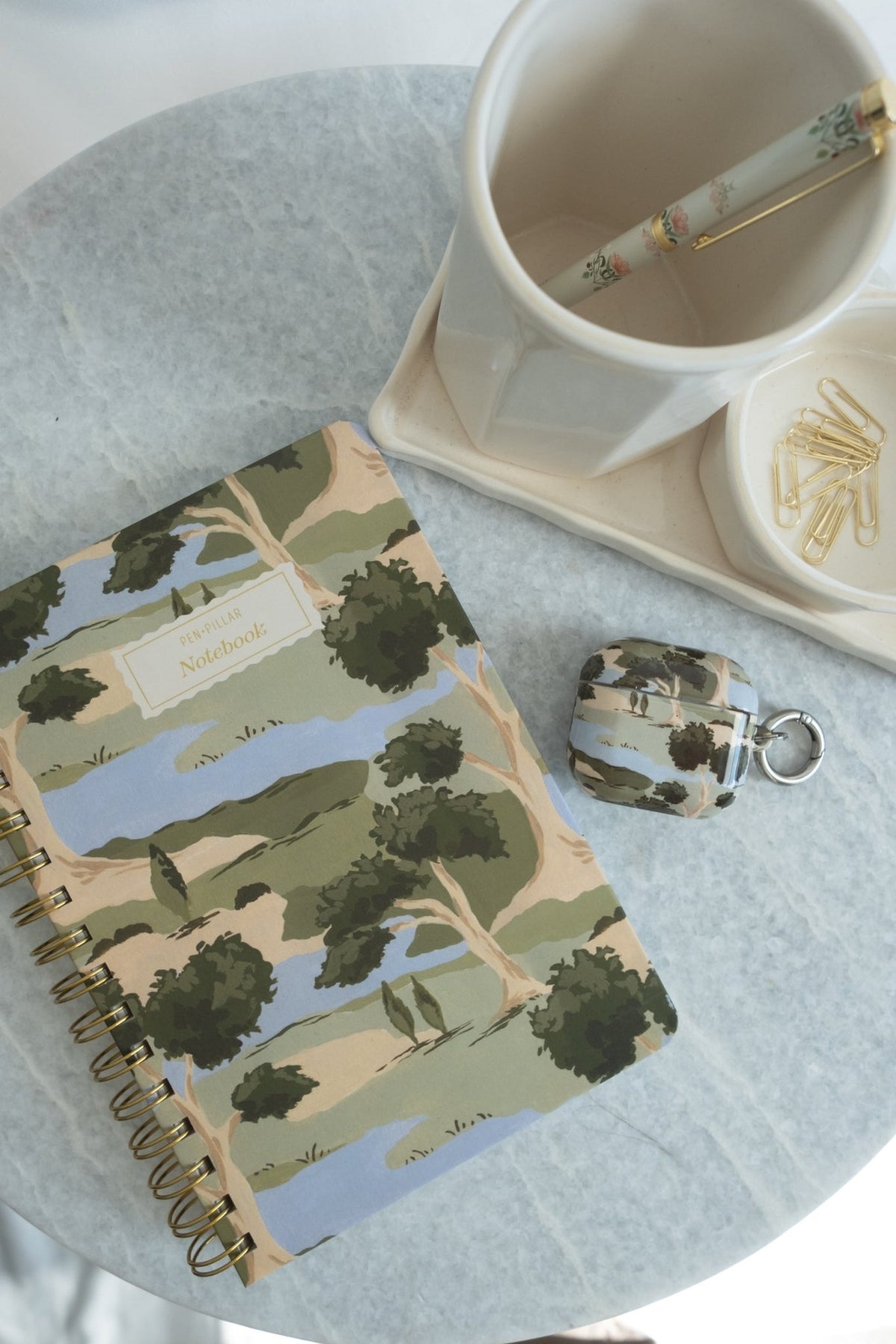 Orchard notebook with a colorful landscape design, placed on a marble table beside a cup and paper clips.