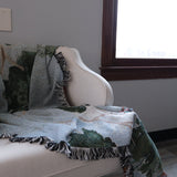 Orchard woven blanket draped over a light-colored chair, featuring green and blue patterns with fringed edges.