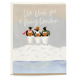 Three penguins wearing scarves and a hat stand on a snowy mound, with "We wish you a Merry Christmas" written above.