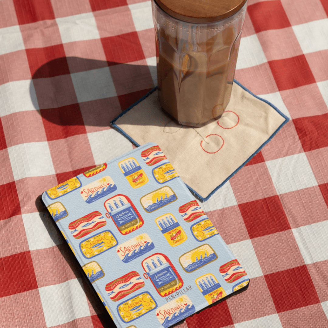 Light blue PEN+PILLAR Kindle case with a colorful sardine tin pattern on a red and white checkered tablecloth.