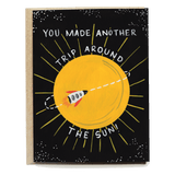 Birthday card featuring a yellow sun and a rocket, with text saying "You made another trip around the sun!" on a black bac...