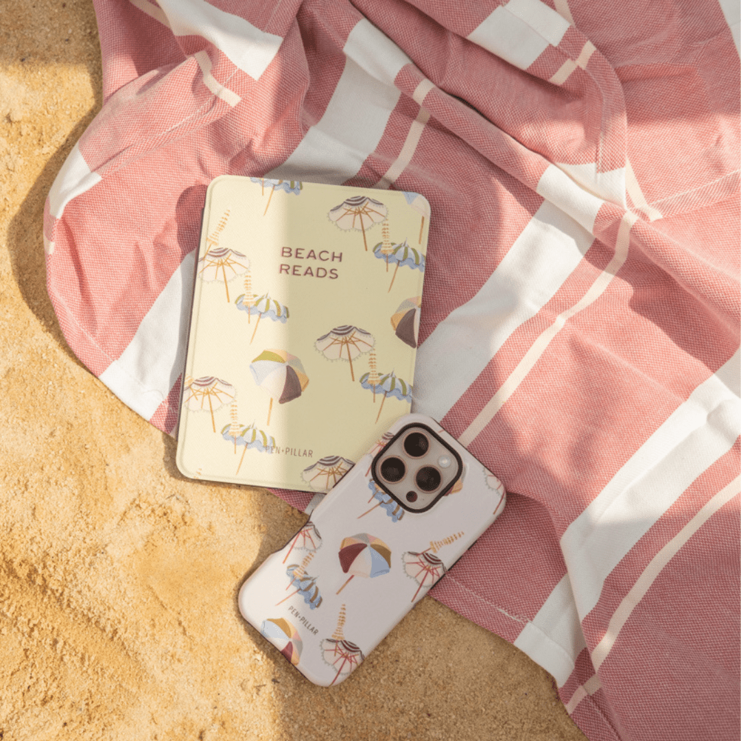White iPhone case and book with umbrella patterns on a pink striped towel and sand. PEN+PILLAR and BEACH READS printed.
