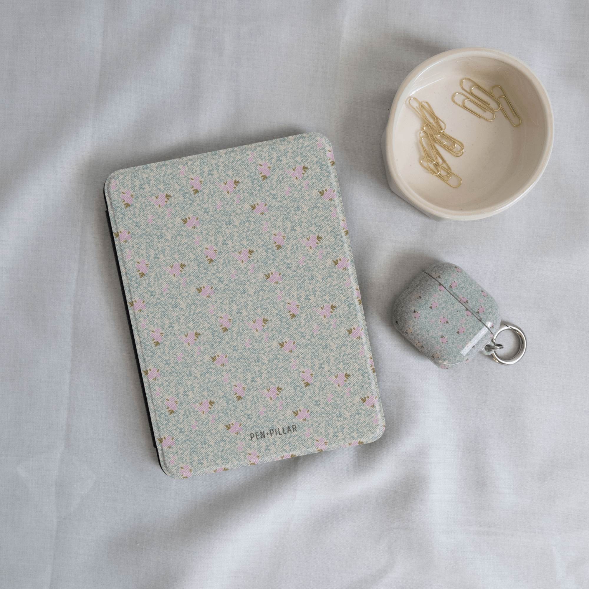 Vine+Bloom Kindle case with a floral pattern in pastel colors, placed on a light fabric surface alongside paper clips.